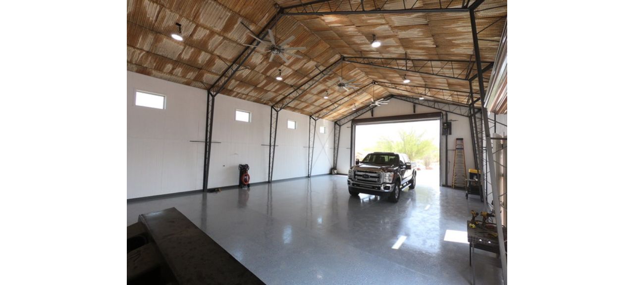 A spacious garage shop with a high ceiling features a large, partially open door. Inside, a pickup truck is parked on the shiny gray floor. The walls have high windows, and there are metal beams and ceiling fans overhead. Natural light fills Breezy Court's impressive space.