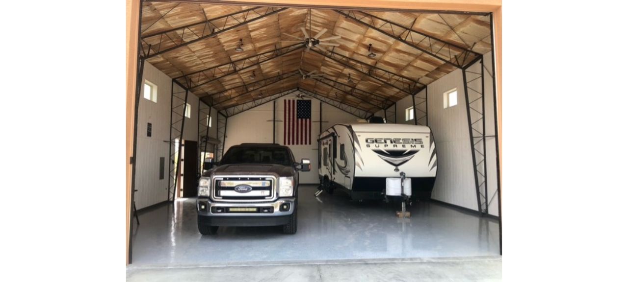 A large garage shop on Breezy Court houses a Ford truck and a Genesis Supreme trailer. The wooden ceiling is supported by black metal beams, and a large American flag proudly hangs on the back wall.