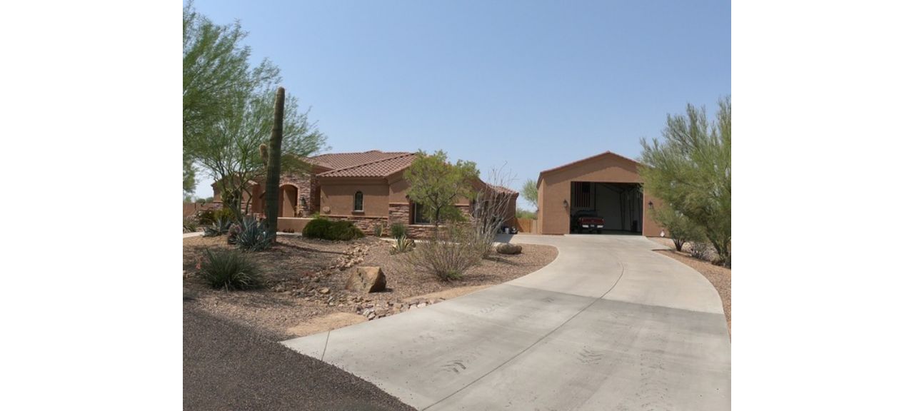 A desert-style home with a stucco exterior and a red-tiled roof sits on Breezy Court. A curved driveway leads to an open garage shop. The landscape features arid plants and rocks under a clear blue sky.