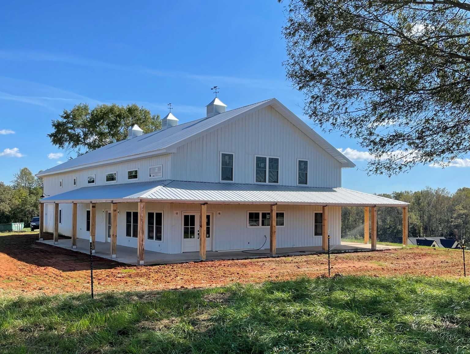 A large white barn-style building with a metal roof stands on a grassy and partially bare soil lot. The structure has a wide porch supported by wooden posts and is surrounded by trees under a clear blue sky.
