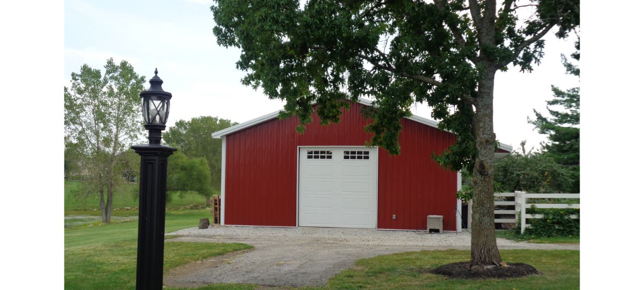 A red barn with a large white door, reminiscent of the Brewster Family Garage, is surrounded by green trees and a grassy lawn. A black vintage-style lamp post stands in the foreground. The sky is overcast.