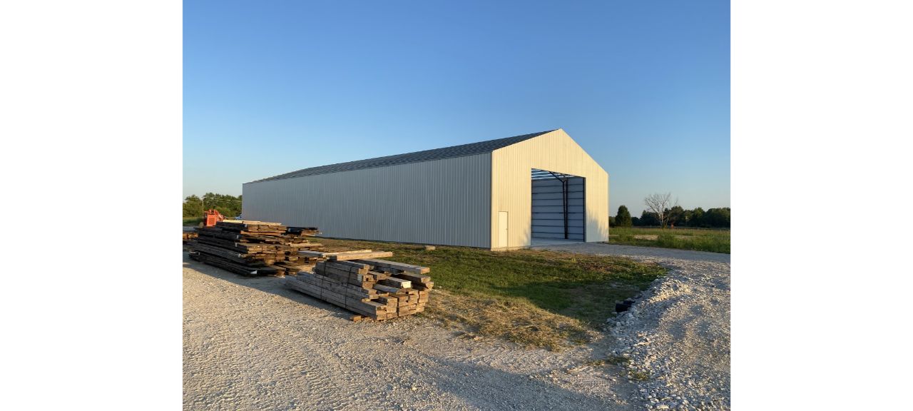 A large, open-ended metal building stands on a gravel path under a clear blue sky. Stacks of reclaimed timber are neatly piled beside it, surrounded by grass and Elmwood trees in the background. The sun casts soft shadows across the scene.