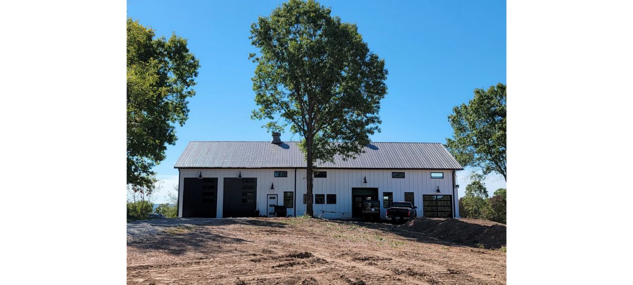 A white barn-style building with a gray metal roof sits on a cleared area, resembling a charming barndo. Large garage doors and windows are visible, flanked by tall trees against the clear blue eye of the sky. A truck is parked outside.