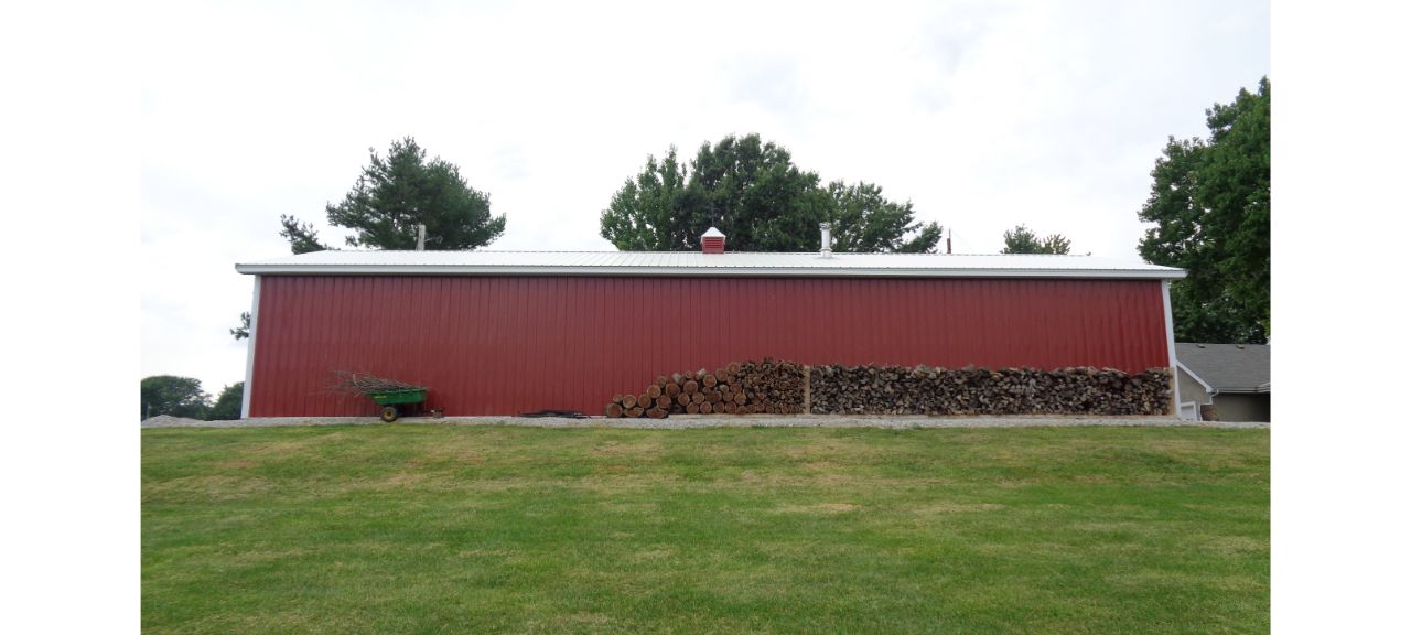 A large red barn with a white roof stands on a grassy hill, reminiscent of the Brewster Family's garage shop. A stack of cut firewood lines the base of the barn, and a green wheelbarrow is parked nearby. Tall trees rise in the background against a cloudy sky.