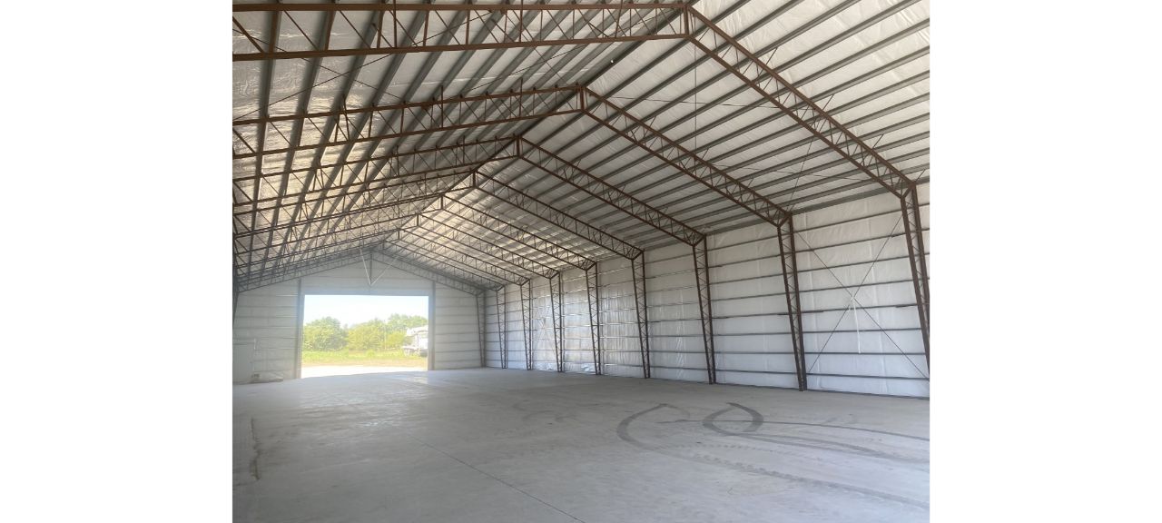 The interior of the large, empty Elmwood warehouse features a high ceiling and open front. Sunlight streams through, illuminating the concrete floor and metal beams, while trees and sky form a picturesque backdrop.
