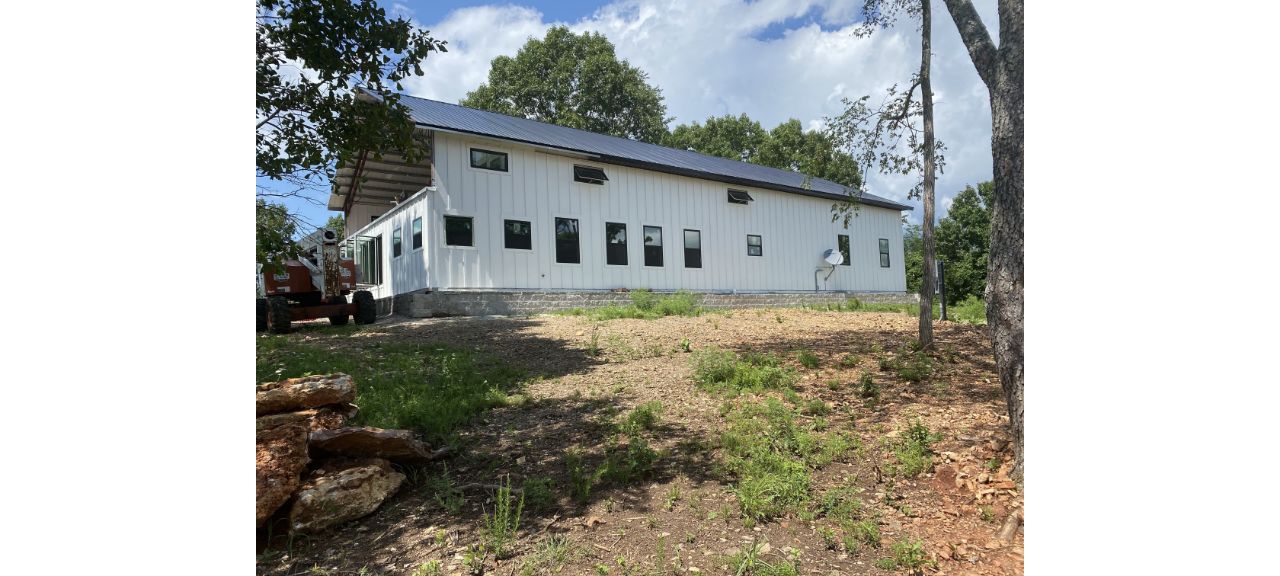 A white metal building with a blue roof reminiscent of a barndo stands on a patch of land. The foreground features scattered grass and rocks, with several trees around, offering a serene Blue Eye charm. The sky is partly cloudy, adding to the tranquil setting.