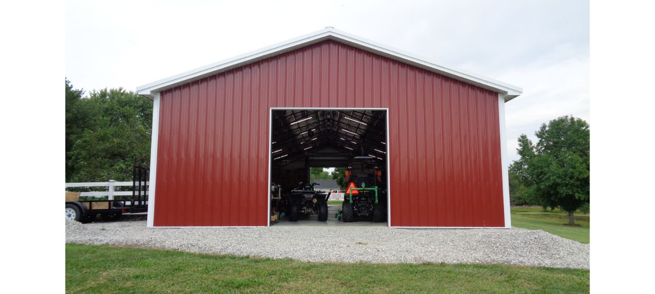 The Brewster Family's large red barn, resembling a vintage garage shop, boasts an open sliding door revealing farm equipment inside. Encircled by grass and gravel under a cloudy sky, trees stand silhouetted in the background.