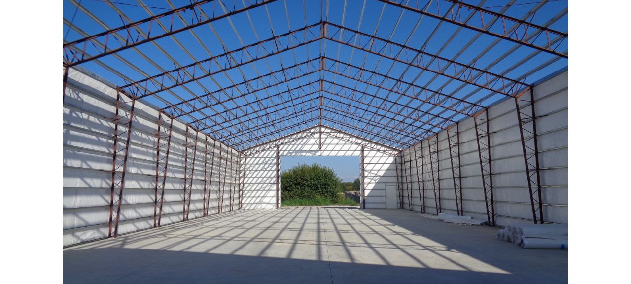Interior view of a large metal-framed warehouse under construction. The roof structure is visible, and the floor is flat concrete. Sunlight streams through the open sides, casting geometric shadows inside. A bush is visible outside the open end, contrasting with reclaimed timber accents.