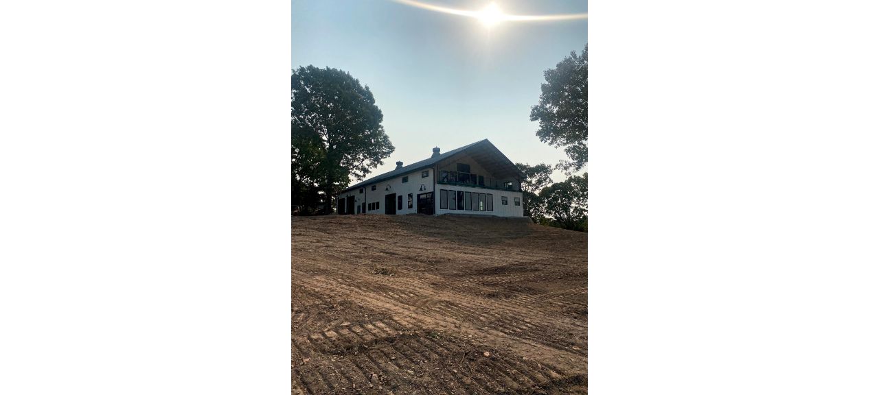 A large two-story Barndo with white walls and a gray roof is nestled on a sloped, freshly tilled dirt plot. Towering trees flank the house, while the sun shines brightly in the sky behind it, casting a serene glow over this charming property.