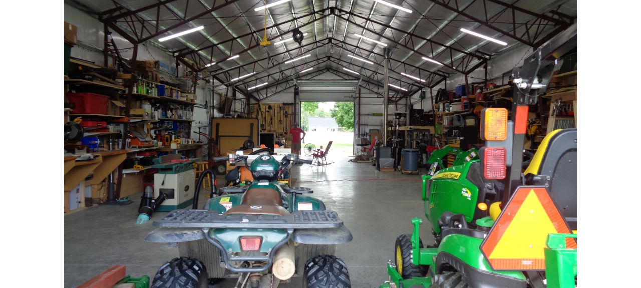 Inside the Brewster Family garage, a spacious haven filled with various tools and equipment, multiple ATVs and a tractor are parked on the concrete floor. Shelves line the walls, holding supplies, while the large open door reveals greenery and light outside.