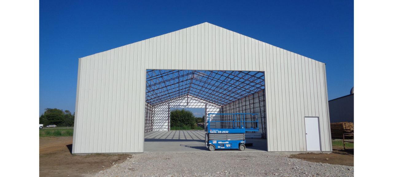 The Elmwood construction site features a large metal building with an open front revealing its skeletal frame. Inside, a blue lift rests on the concrete floor. The structure, surrounded by a clear blue sky and grassy area, hints at the use of reclaimed timber for future additions.