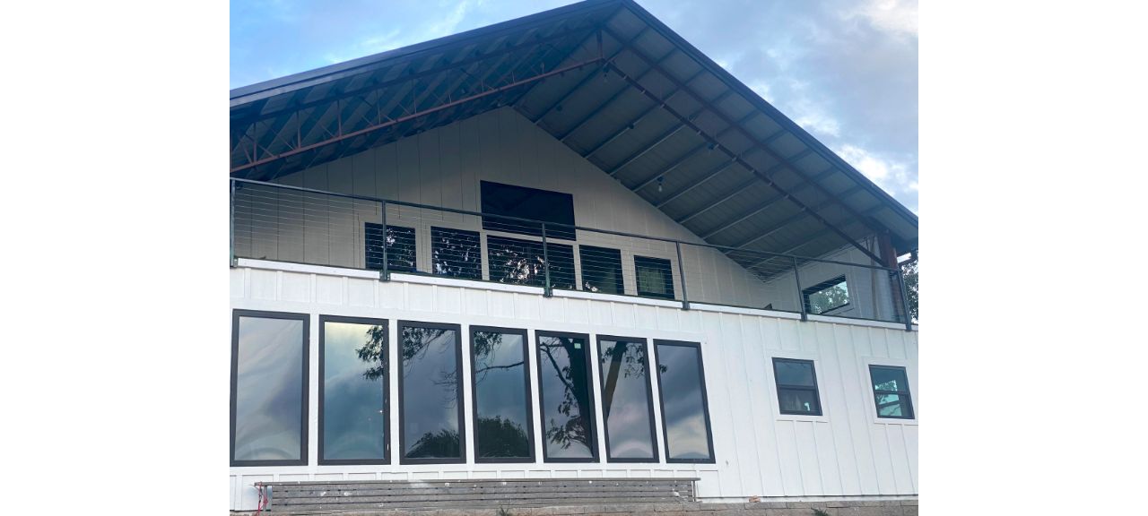 A modern, two-story lakehouse features large glass windows on the lower level and a sloped roof. The upper level has a balcony with a metal railing, offering serene views of Blue Eye. The sky is partially cloudy, adding to the tranquil ambiance.