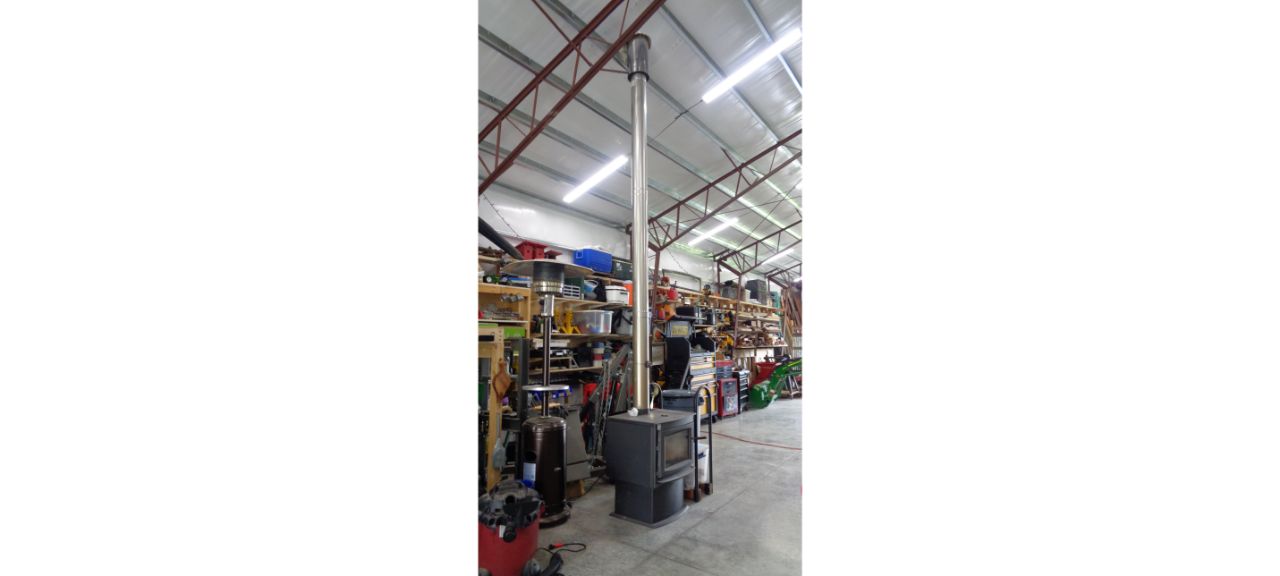 A large, vertical metal stove with a long chimney stands proudly in the spacious Brewster Family Garage. The background features shelves filled with various tools and equipment, under a high ceiling illuminated by fluorescent lighting.