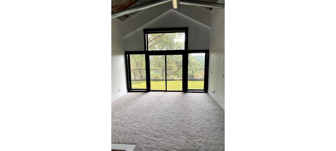 A room with high vaulted ceilings and large black-framed windows overlooking a lawn and trees, evoking a serene lakehouse feel. The space is carpeted, with natural light streaming in from outside, creating an inviting Blue Eye ambiance.