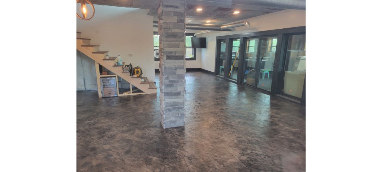 A spacious, modern lakehouse basement with polished concrete floors. A staircase with wooden steps is on the left, a stone pillar in the center, and large glass doors line the right side. The space is brightly lit, with some tools and a TV visible.
