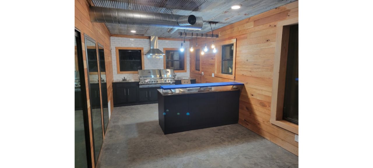 A modern kitchen interior reminiscent of a chic barndo, featuring wooden walls, concrete flooring, and a corrugated metal ceiling. The space includes a sleek black kitchen island with a glossy surface, a stainless steel range hood, pendant lights, and large windows.