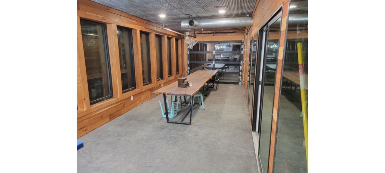 A long wooden room akin to a lakehouse, with large windows on the left side and a lengthy wooden table with metal legs in the center. The cement floor complements shelves at the far end, while hanging lights illuminate this serene escape.