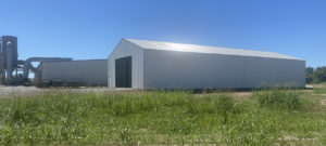 A large, white industrial building with a peaked roof stands in a grassy area under a clear blue sky. Nearby, another similar Elmwood structure features visible ventilation equipment, adding to the harmonious landscape.