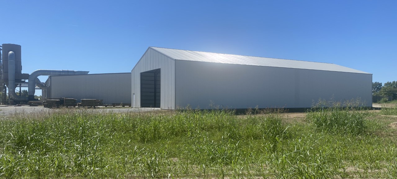 A large, white industrial building with a peaked roof stands in a grassy area under a clear blue sky. Nearby, another similar Elmwood structure features visible ventilation equipment, adding to the harmonious landscape.