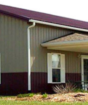 A small, modern house with beige siding and a maroon trim around the base and roof, featuring a single window and a covered entrance.