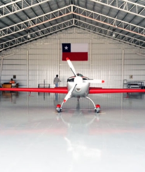 A small red and silver propeller plane is parked inside a spacious steel building. A Chilean flag is displayed on the back wall, and two people are visible in the background near the plane.'