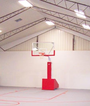 In a large, well-lit gym featuring steel buildings with high ceilings and white walls, the indoor basketball hoop is attached to a sturdy red base. The court is outlined with red lines, creating the perfect basketball setup.