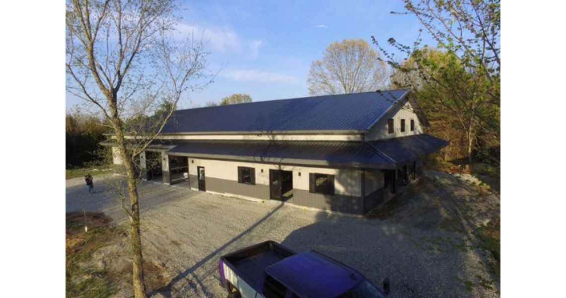 An aerial view of a modern barn-style building in Buxton showcases a dark metal roof and light-colored walls. Surrounded by trees, it features a gravel driveway leading to the poolhouse, with a vehicle parked in the foreground.