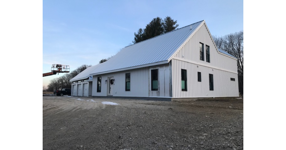A sleek, white barndominium with a metal roof, large windows, and several garage doors. It sits on a dirt clearing surrounded by trees under a clear sky.
