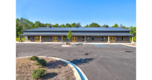 A single-story building with a dark roof and beige walls, the Kudzu Staffing Office is situated in a parking lot with marked spaces, including accessible parking. The background features green trees and a clear blue sky.