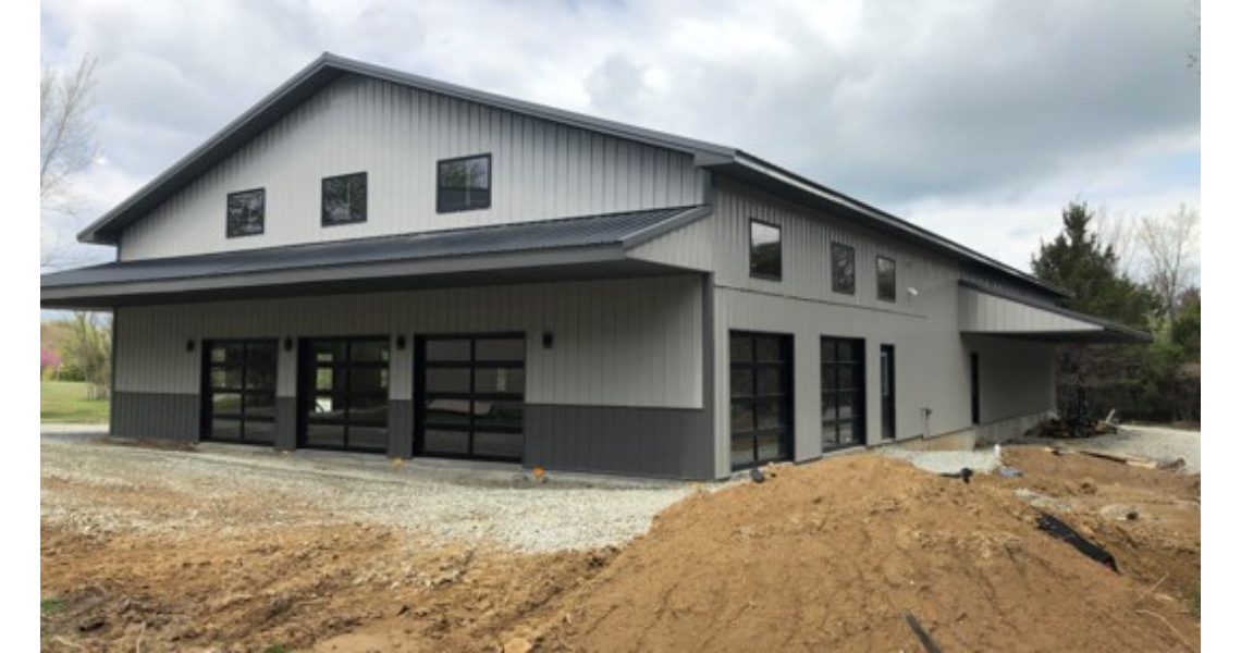 A modern, gray metal building resembling a poolhouse with large windows and garage doors sits on a plot of gravel and dirt. The cloudy sky looms over Buxton, with trees in the background adding a touch of nature.