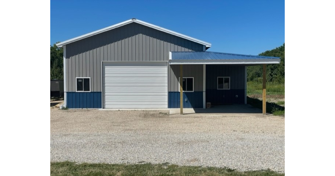 A blue and gray metal garage featuring a large white door and an attached covered area on the right, sits prominently on a gravel driveway. With grass and trees in the background under Bonham's clear blue sky, this building is as functional as it is picturesque.