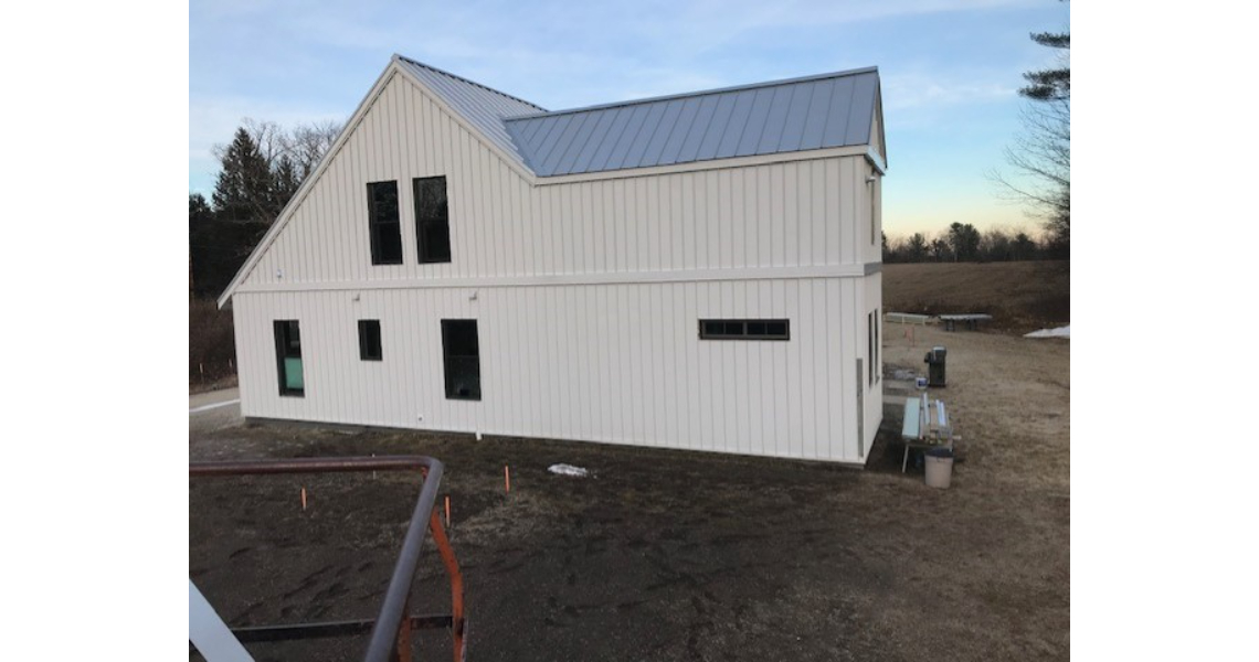 A white barndominium with a metal roof, nestled in a rural setting. This Malm-inspired house features multiple large windows, surrounded by a grassy field and distant trees. The sky is clear with a hint of blue.