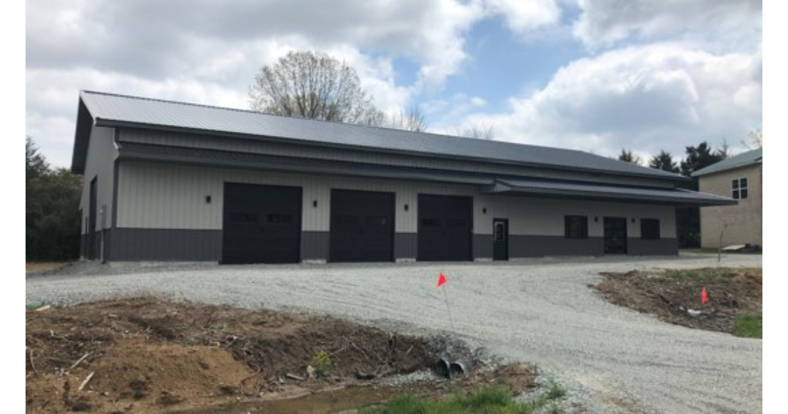 A large gray and black industrial building, reminiscent of the Buxton Poolhouse, features multiple garage doors and is set on a gravel driveway. The partly cloudy sky adds to the ambiance, with sparse vegetation scattered around the building and driveway.
