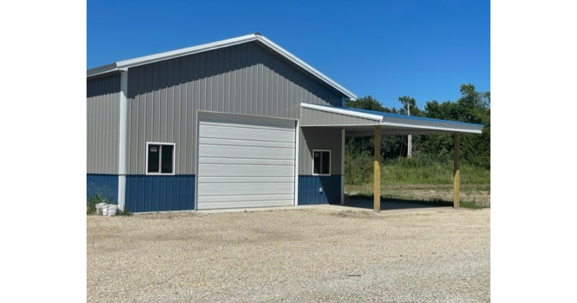 A Bonham metal garage with a small awning, two windows, and a large closed garage door stands on a gravel lot. The shop is gray with a blue base, set against a backdrop of trees and a clear blue sky.