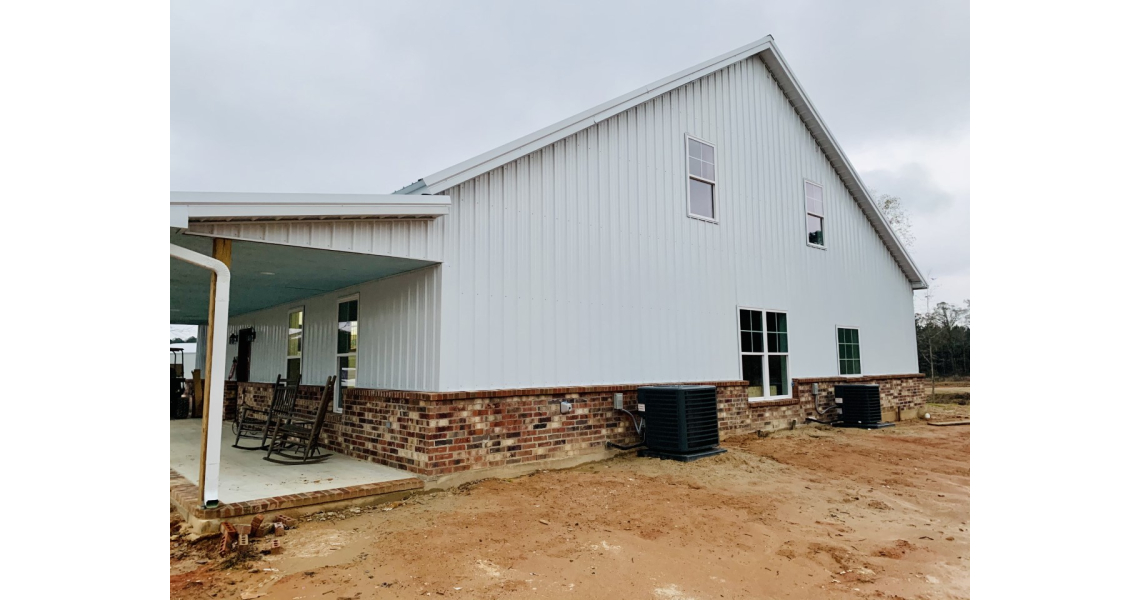 A large, white metal building with a brick base is shown, resembling a classic barndominium. It features several windows, with two air conditioning units visible outside and a covered patio with rocking chairs on the left side. The overcast sky looms above the bare soil ground.