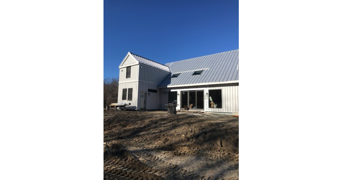 A modern, light gray two-story house with a metal roof resembles a sleek barndominium. Large windows reflect the clear blue sky, while the bare ground suggests recent construction or landscaping work.