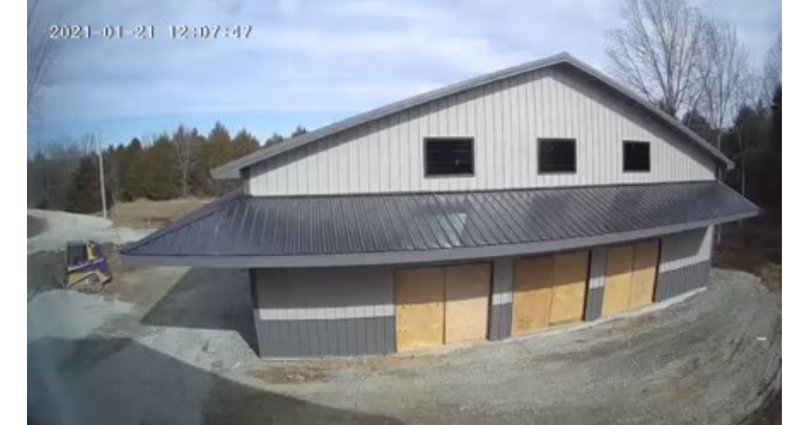 A new barn-like poolhouse with a metal roof and boarded windows sits in a rural Buxton area, surrounded by trees. The gravelly ground adds to its rustic charm. A timestamp reads 