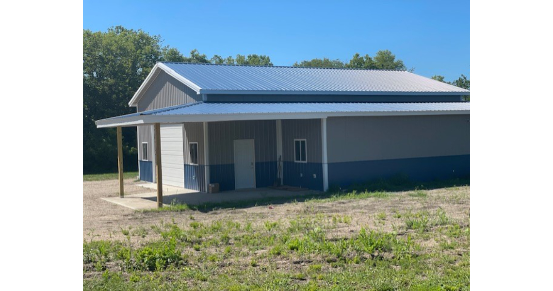 A small, single-story Bonham Garage with a blue and gray exterior and a metal roof. It features a porch supported by wooden posts. The building is set in a grassy area with trees in the background under a clear blue sky, offering both charm and functionality.