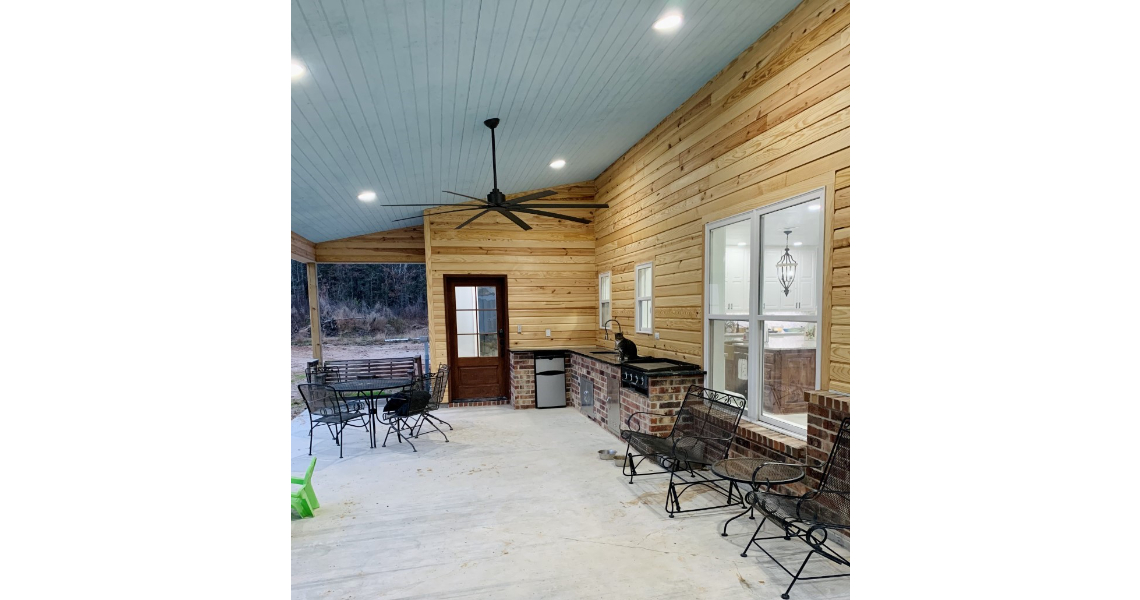 The covered patio, reminiscent of a Chapman barndominium, features wooden walls and a ceiling fan. A glass door and windows line one wall, while a grill and metal seating area rest on the concrete floor. The surrounding outdoor scenery creates a natural backdrop.