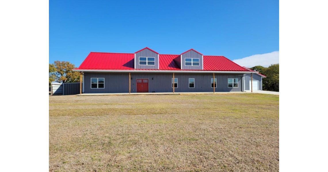 The Freeman Residential Home is a charming single-story house with a gray exterior and a vibrant red roof. It features two dormer windows and a brown double front door, nestled under the clear blue sky, surrounded by a grassy yard and some trees.