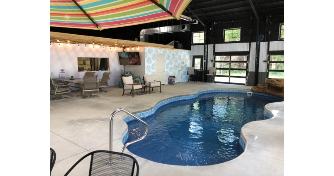 The Buxton Poolhouse boasts an indoor area with a charming freeform swimming pool, surrounded by a concrete deck. Relax on lounge chairs or dine at the table under a colorful striped umbrella. The ambiance is enhanced by patterned wallpaper and twinkling string lights.
