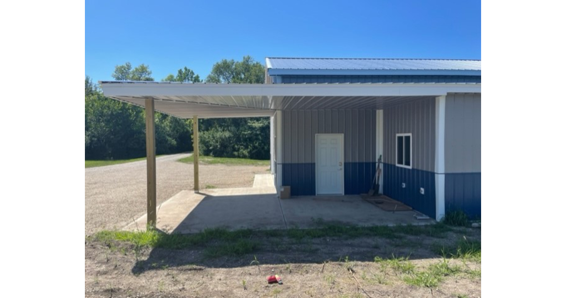 A metal building in Bonham with a white and blue exterior features a covered carport supported by wooden beams. This garage is set on a concrete slab, surrounded by grass and gravel, with trees in the background under a clear blue sky.