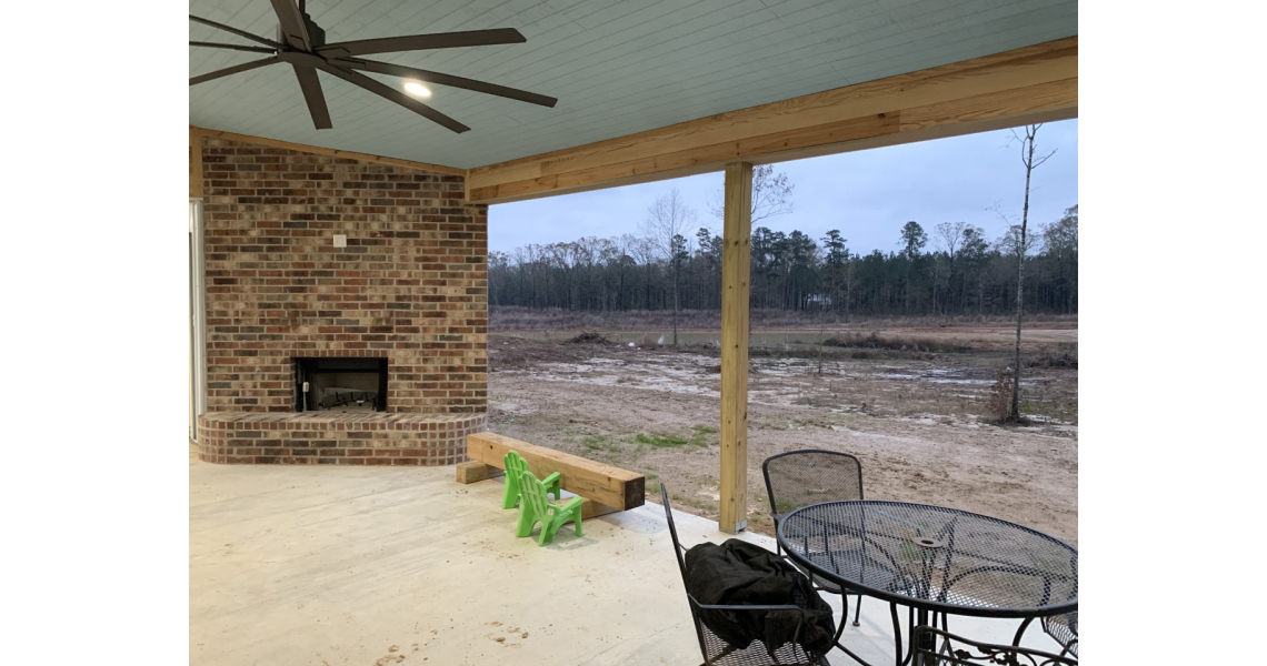 A covered patio in a Chapman-style barndominium features a brick fireplace, ceiling fan, and metal table with chairs. Two green children's chairs face the fireplace, while the view includes a grassy field and trees under a cloudy sky.