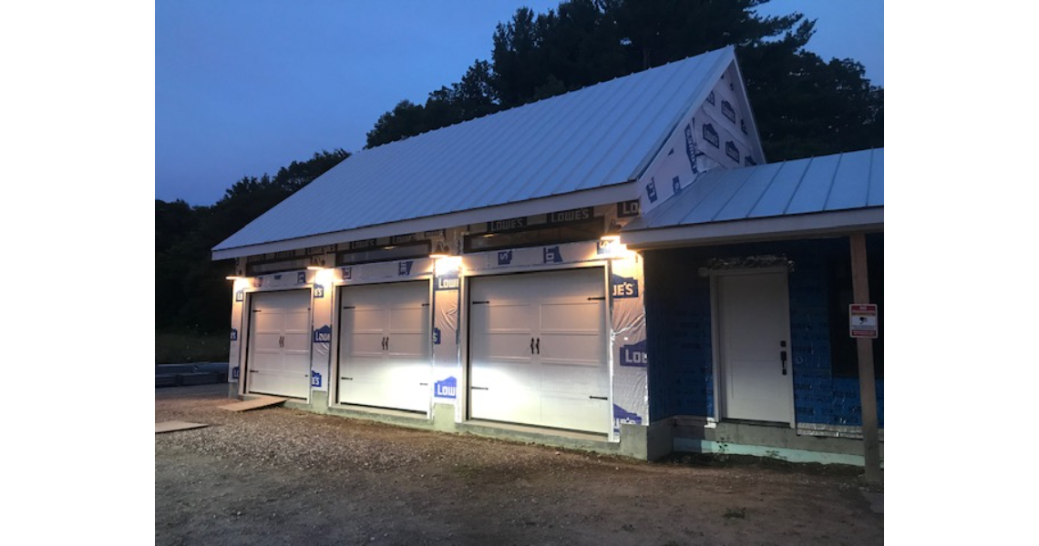 A barndominium under construction features three illuminated garage doors at dusk. The structure boasts a white metal roof and partially completed exterior walls with visible construction materials and logos. Malm trees stand gracefully in the background, adding to the picturesque setting.