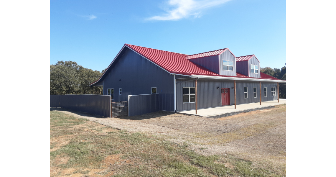 A modern barn-style home with gray walls and a red roof, featuring a large sliding door and several windows. Set against a clear blue sky, it embodies the charm of Freeman Residential living, surrounded by lush grass and trees in the background.
