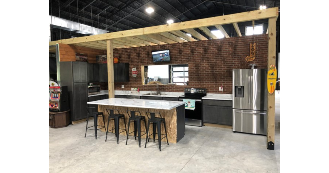 A modern kitchen setup in an open space reminiscent of Buxton Poolhouse, with brick walls, a large island with four barstools, dark cabinets, stainless steel appliances, and a small TV mounted on the wall. A wooden beam ceiling elegantly frames the kitchen area.