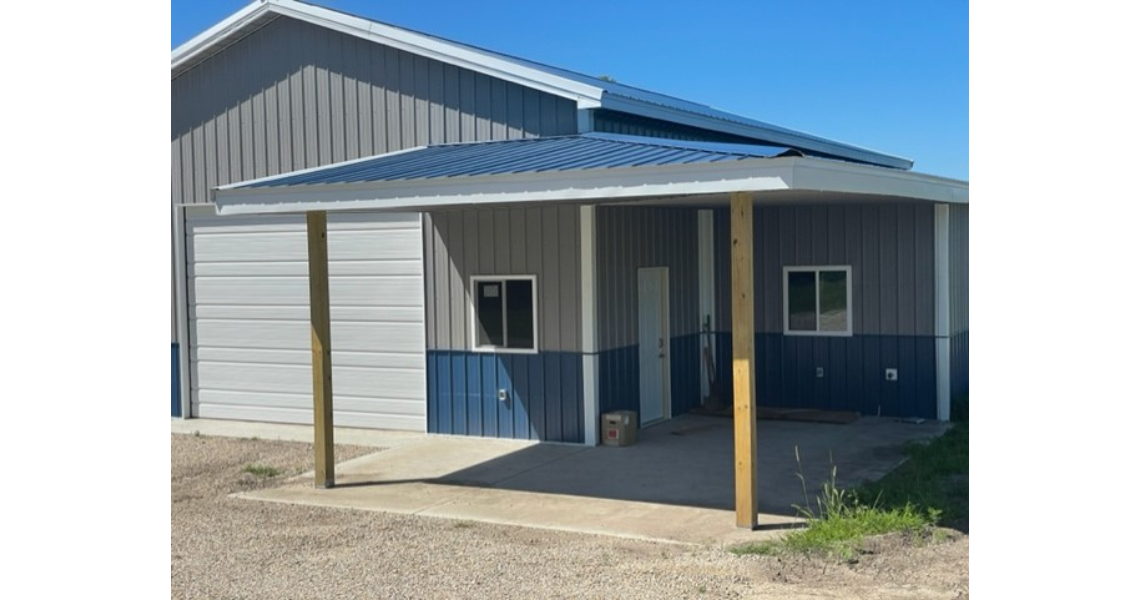 A large gray and blue metal shop with a white garage door and a covered porch stands proudly in Bonham. The porch, supported by two wooden beams, features a blue metal roof. This garage-like structure rests on a concrete foundation with patches of grass visible nearby.