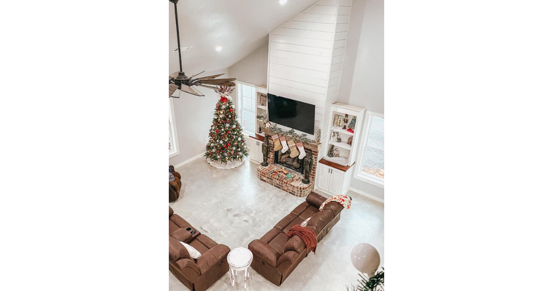 A cozy living room in a Chapman-style Barndominium features a vaulted ceiling and a decorated Christmas tree beside a stone fireplace adorned with stockings. Two brown sofas face the fireplace, with a ceiling fan gently rotating above.