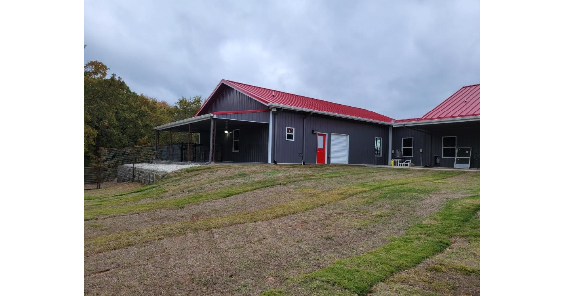 The Freeman Residential building, with its dark exterior and striking red metal roof, sits gracefully on a grassy hill. Featuring a red door, several windows, and a covered porch area, this modern residential home stands out against the overcast sky.