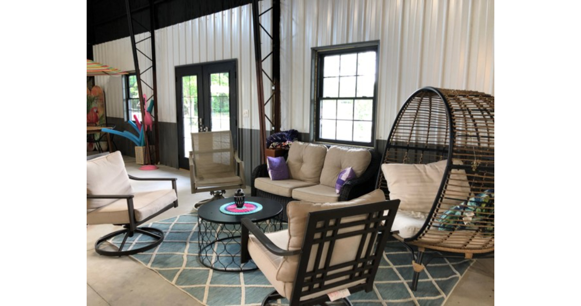 A cozy indoor poolhouse patio area features a wicker egg chair, a beige cushioned loveseat, and two matching armchairs. A round coffee table sits centered on a blue geometric rug. Black-framed windows and doors offer a Buxton-like view of the greenery outside.
